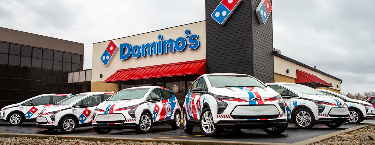 A row of Domino's-branded electric cars with red, white, and blue wraps parked in front of a Domino's restaurant.